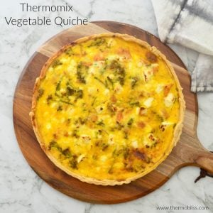 A vegetable quiche with a pastry crust resting on a round wooden serving board.