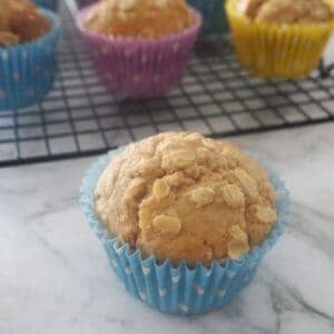 A banana and oat muffin in a blue muffin case in front of a wire rack filled with more muffins cooling