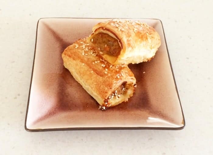 Two golden baked sausage rolls sprinkled with sesame seeds in a square bowl