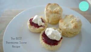 Plain scones on a plate with one scone cut in half and topped with jam and whipped cream.
