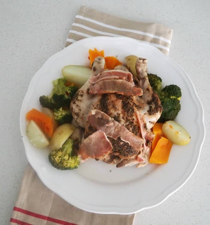 A serving plate with a roasted chicken coated with herbs and bacon, surrounded by cooked potatoes, carrots and broccoli.
