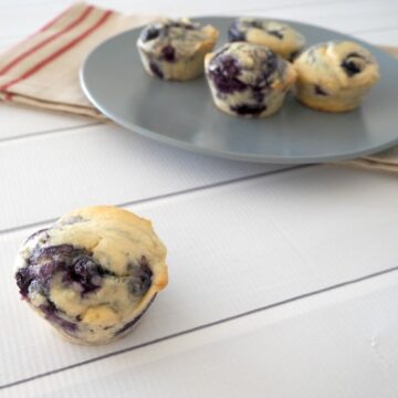 Four muffins baked with blueberries on a plate in the background, with one on the bench in the foreground.