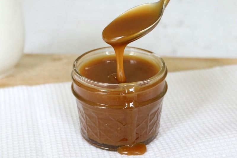 A small glass jar filled with caramel sauce, with a spoon held above drizzling caramel sauce into the jar.
