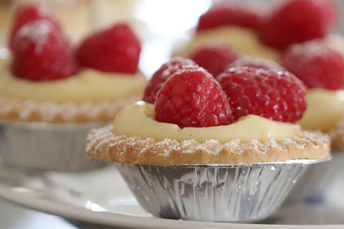 A close up of a mini custard tart in a silver tart case, with fresh raspberries on top.