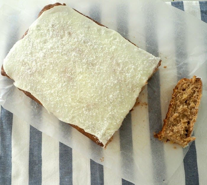 An overhead shot of a rectangular loaf made with banana and pineapple, and white icing, and one slice cut and resting beside.