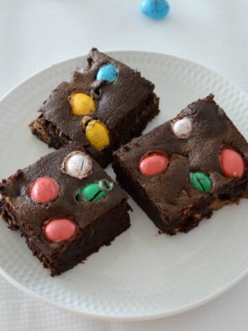 Three pieces of a chocolate brownie slice with coloured Easter eggs baked on the top, served on a white plate