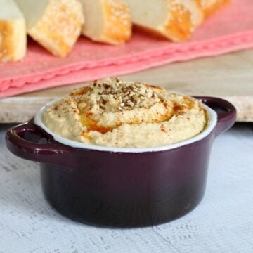 A ramekin filled with hommus dip, with bread stick slices in the background