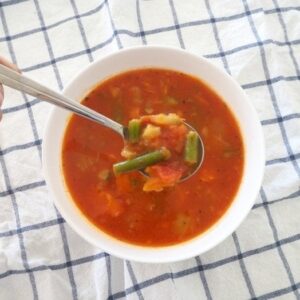 A spoonful in a bowl of red tomato and chunky vegetable soup, on a blue and white striped tea towel.