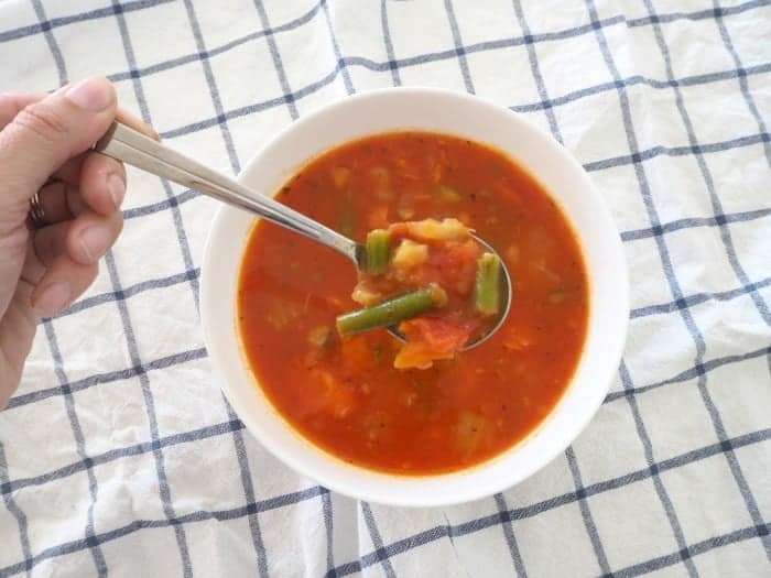 A spoon full of vegetable chunks being lifted out of a bowl of tomato and vegetable soup.