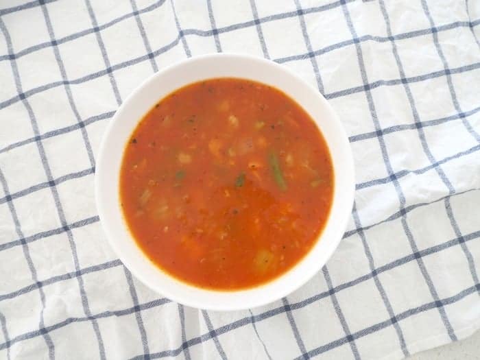 An overhead shot of a bowl of tomato and vegetable soup sitting on a white and blue check tea towel.