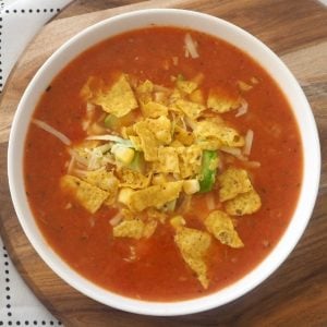 An overhead shot of a bowl of red soup with shredded chicken,corn chips, and grated cheese on top