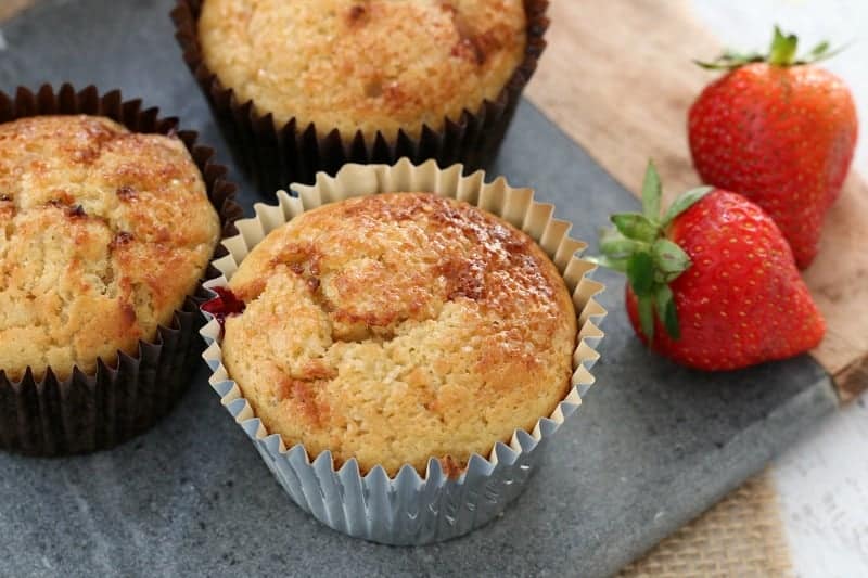Muffins in muffin cases sitting on a marble board with fresh strawberries beside.