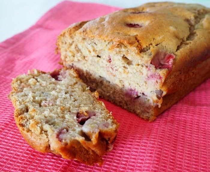 A loaf made with bananas and strawberries with one slice cut, sitting on a pink tea towel.