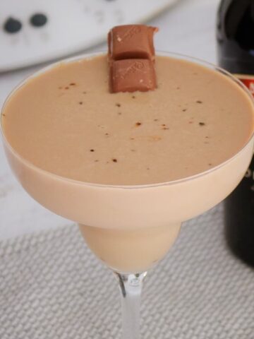 A stemmed cocktail glass filled with a creamy chocolate drink, with a piece of chocolate on the side.