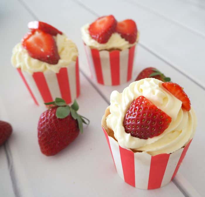 Cupcakes in stripey cases, topped with cream and pieces of fresh strawberries.