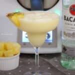 A piece of pineapple on the edge of a creamy cocktail in a stemmed cocktail glass, with a bottle of Bacardi next to it.