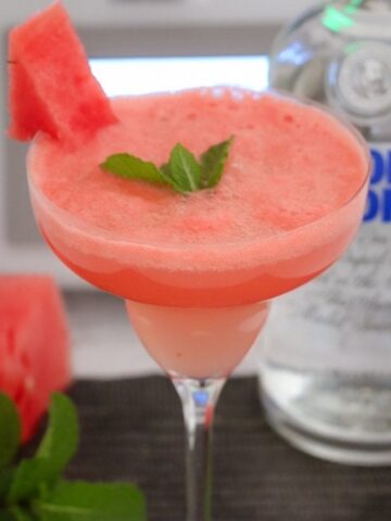 A stemmed cocktail glass with a slushy pink drink garnished with a piece of watermelon and a sprig of mint.