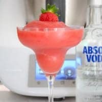 A slushy strawberry drink served in a stemmed cocktail glass with a strawberry on top, and a bottle of vodka nearby.