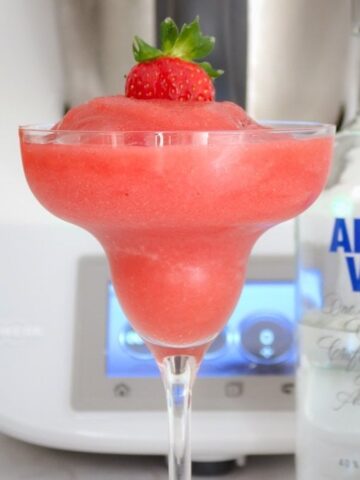 A slushy strawberry drink served in a stemmed cocktail glass with a strawberry on top, and a bottle of vodka nearby.