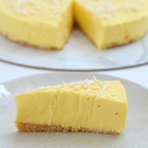 A serve of a creamy cheesecake made with mango on a plate, with the remaining cheesecake in the background.