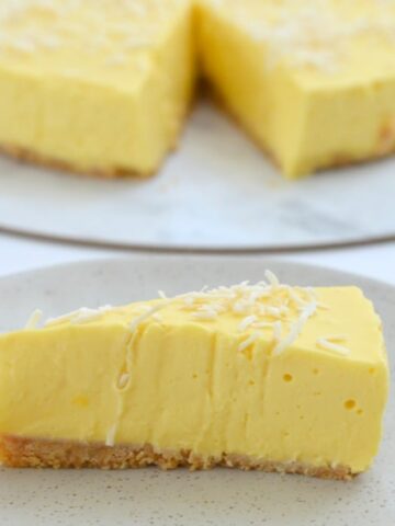 A serve of a creamy cheesecake made with mango on a plate, with the remaining cheesecake in the background.