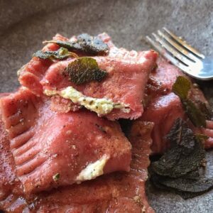 A close up of beetroot coloured ravioli with a goats cheese filling and sage leaves in a bowl.