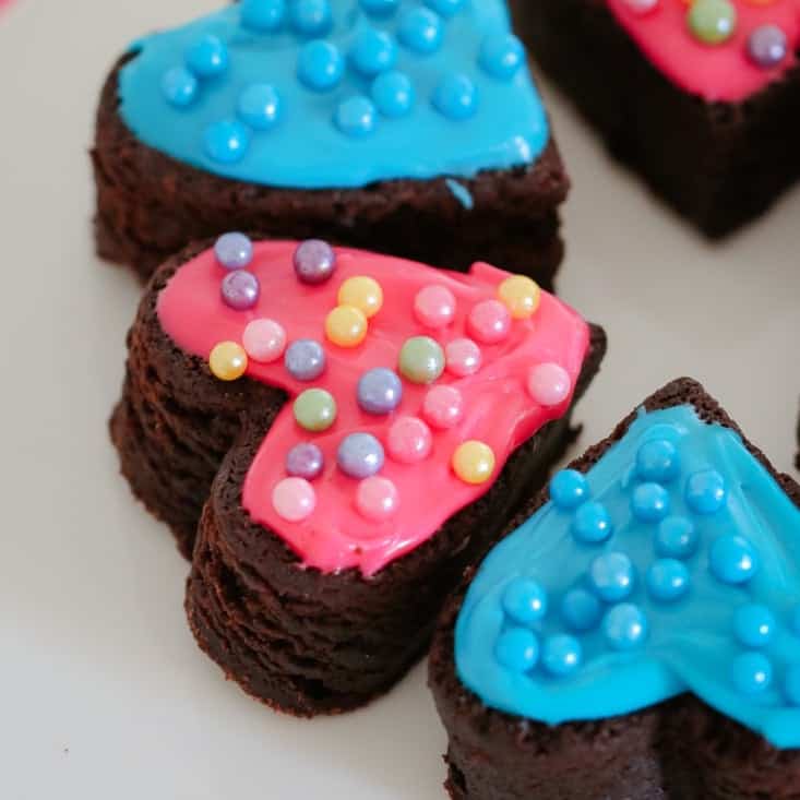 Pieces of a chocolate brownie slice, cut into heart shapes and decorated with pink and blue icing and coloured cachous balls