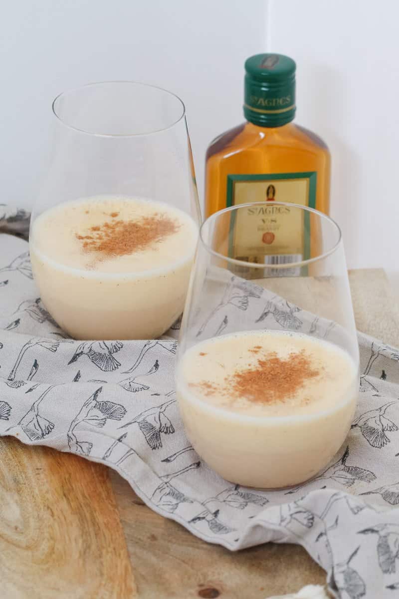 A bottle of brandy behind two glasses of nutmeg topped eggnog.