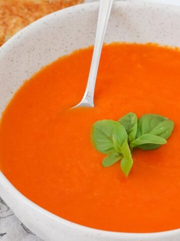 A bowl with a spoon, filled with a creamy red tomato soup and a sprig of basil on top.