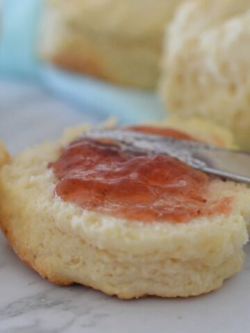 A close up of a knife spreading jam over a scone cut in half.