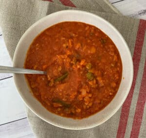 A bowl of red Minestrone soup with a spoon in it, with beans, pasta and vegetables in the soup