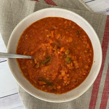 A bowl of red Minestrone soup with a spoon in it, with beans, pasta and vegetables in the soup