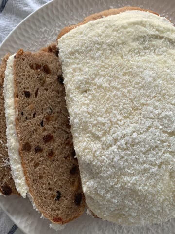 A round Boston Bun, iced and served on a plate, with two slices cut showing sultanas inside.
