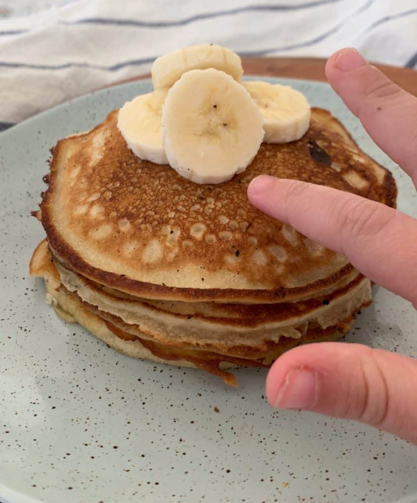 A hand reaching out to a stack of pancakes with slices of banana on top