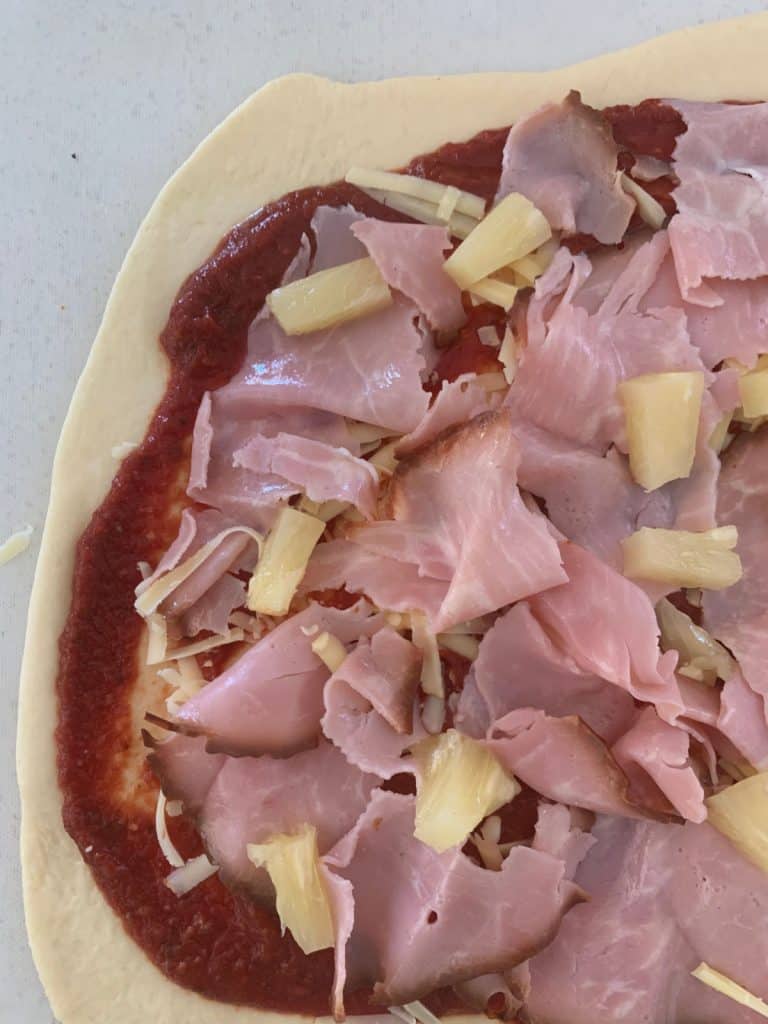 A close up of pizza dough with tomato paste, shaved ham and pineapple on top. 