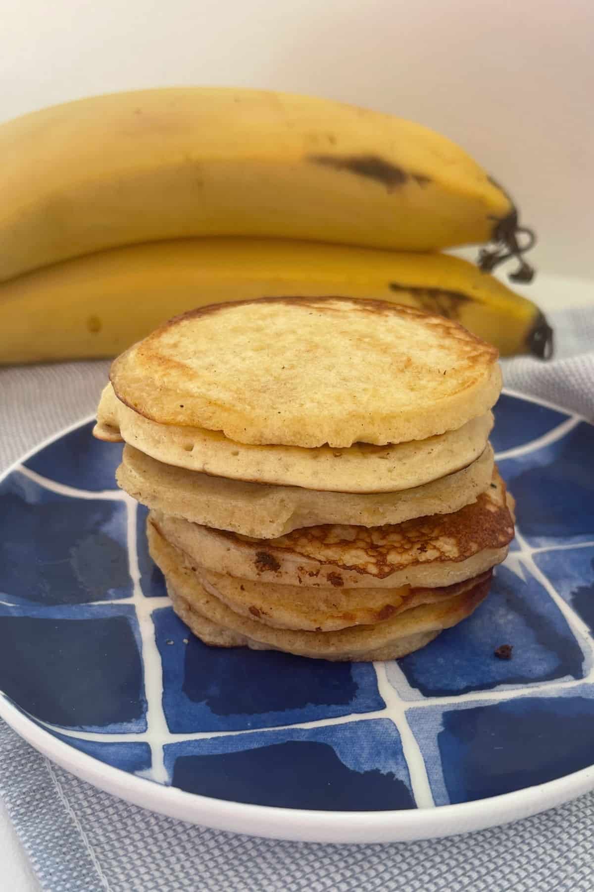 Stack of banana PIKELETS on a blue and white striped plate. In the background there are a bunch of bananas.