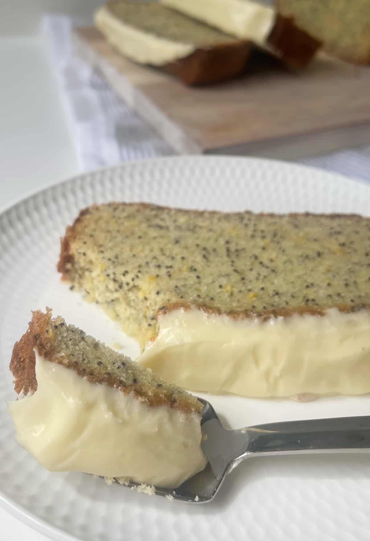 Fork holding a piece of lemon and poppy seed cake. in the background there is a slice of cake on plate.