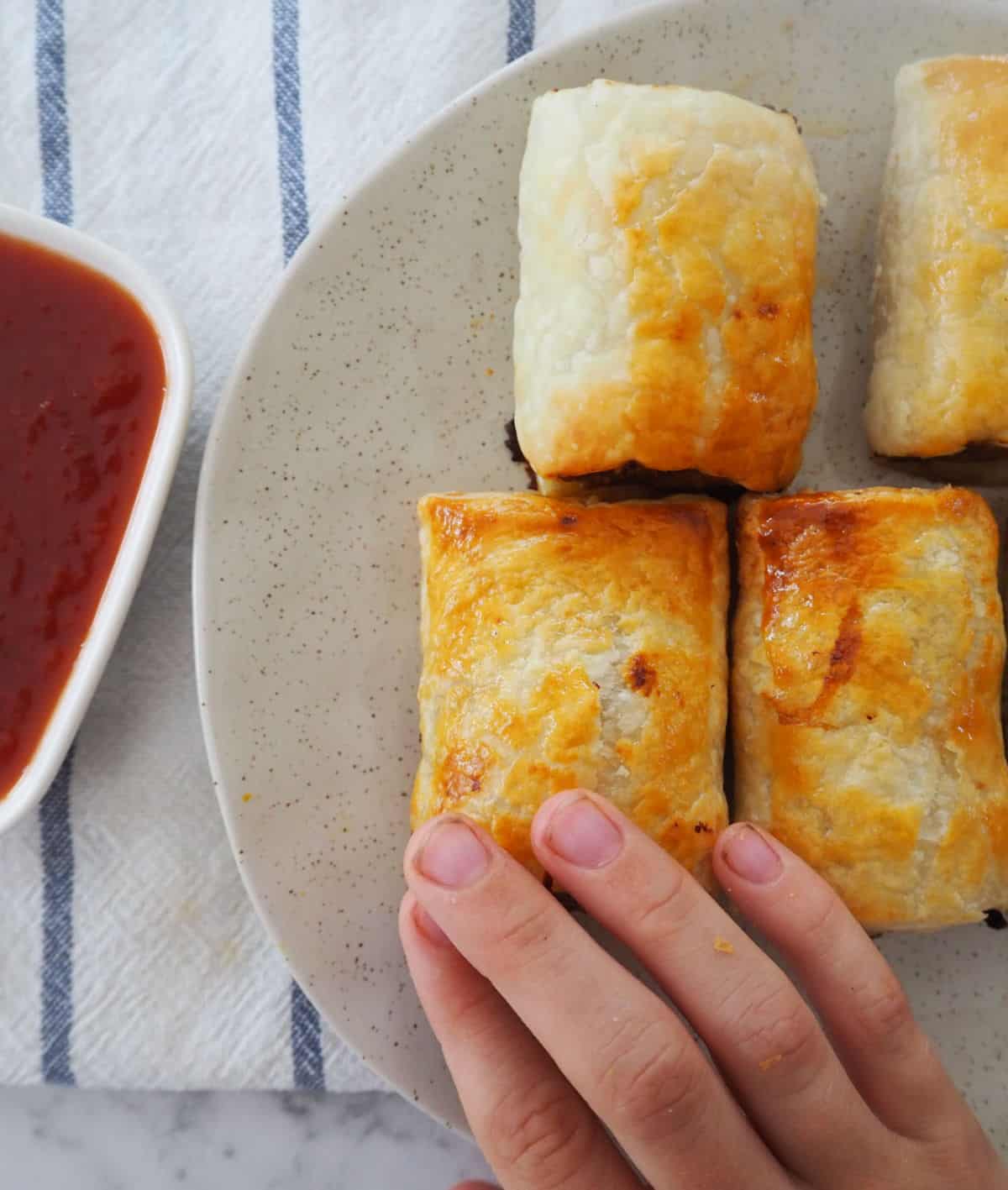 Child's hand reaching for sausage roll on a plate