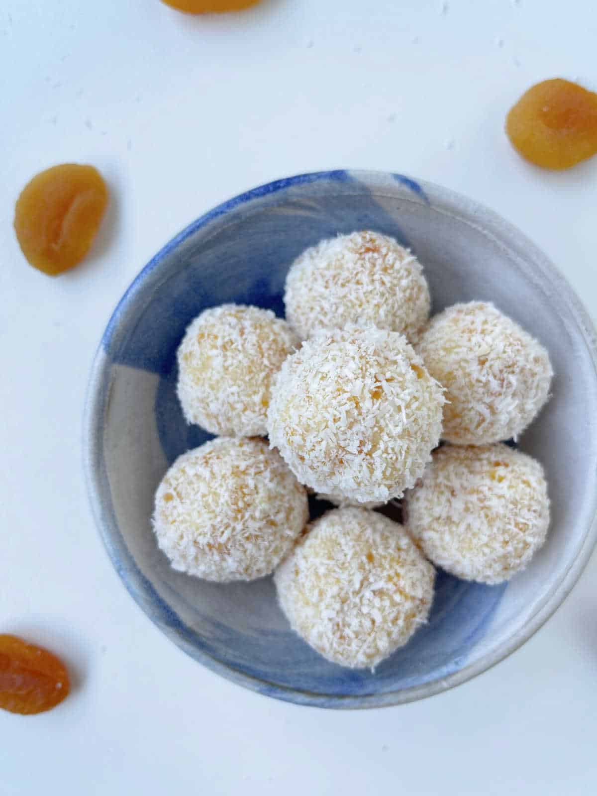 Apricot and coconut balls in a blue and cream bowl with apricots in the background.