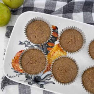 Overhead view of Apple and Cinnamon Muffins sitting on a flowered serving tray. In the background there are green apples and a checkered tea towel.