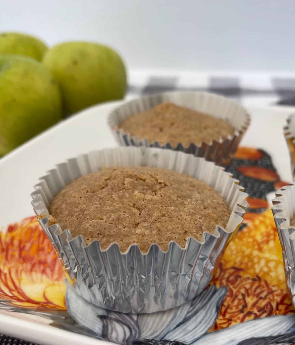 Apple and Cinnamon Muffins sitting on a flowered serving tray. In the background there are green apples and a checkered tea towel.