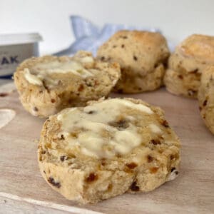 Date scone sliced in half and topped with butter sitting on a pale wooden serving board. In the background there are more Date Scones and a tub of Lurpack butter.