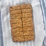 Anzac Slice cut into 16 pieces. Sitting on a sheet of baking paper on top of a blue striped tea towel.