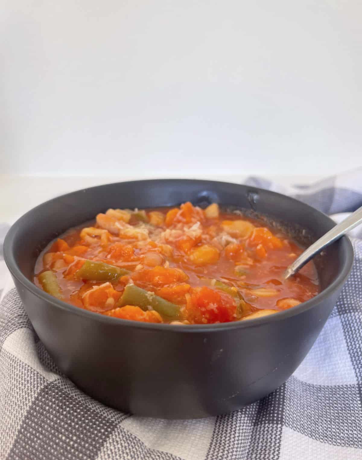 Side view of a black bowl filled with Minestrone Soup that has been made in a Thermomix. There is a silver spoon in the bowl.