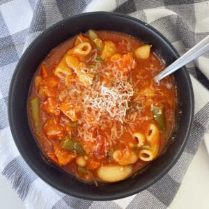 Overhead view of Minestrone Soup in a black bowl that has been topped with grated Parmesan cheese. The bowl is sitting on a black and white checkered tea towel.