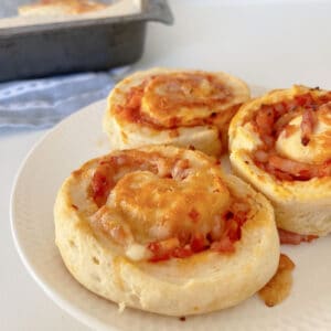 Side view of three pizza scrolls sitting on a white plate. In the background there is a baking tray sitting on a blue striped tea towel.