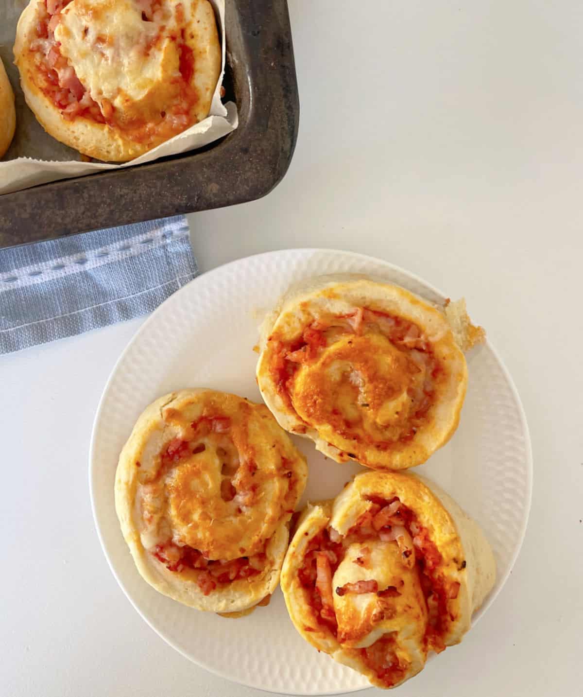 Overhead view of three pizza scrolls sitting on a white plate. In the background there is a baking tray sitting on a blue striped tea towel.