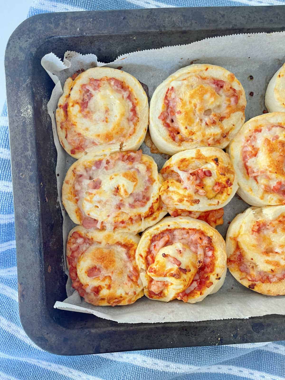 Pizza Scrolls straight from the oven sitting in baking tray which is on top of a blue and white striped tea towel.