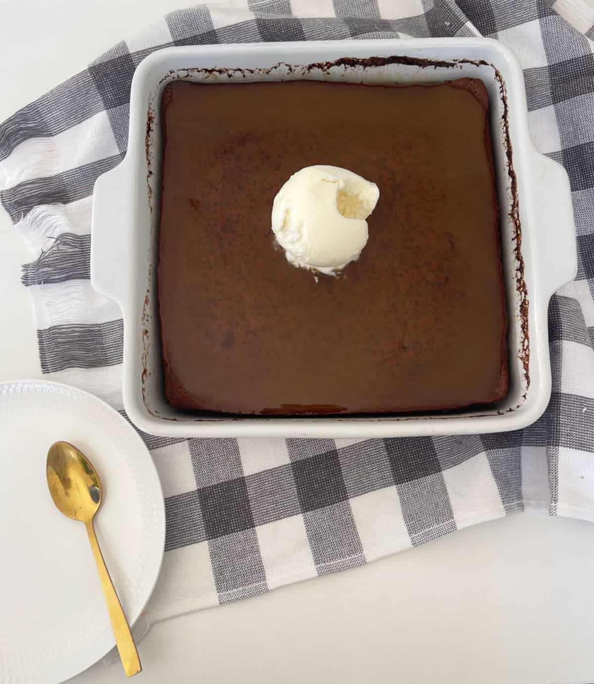 Overhead view of Sticky date pudding in a white square baking dish. A white plate and gold spoon is next to it.
