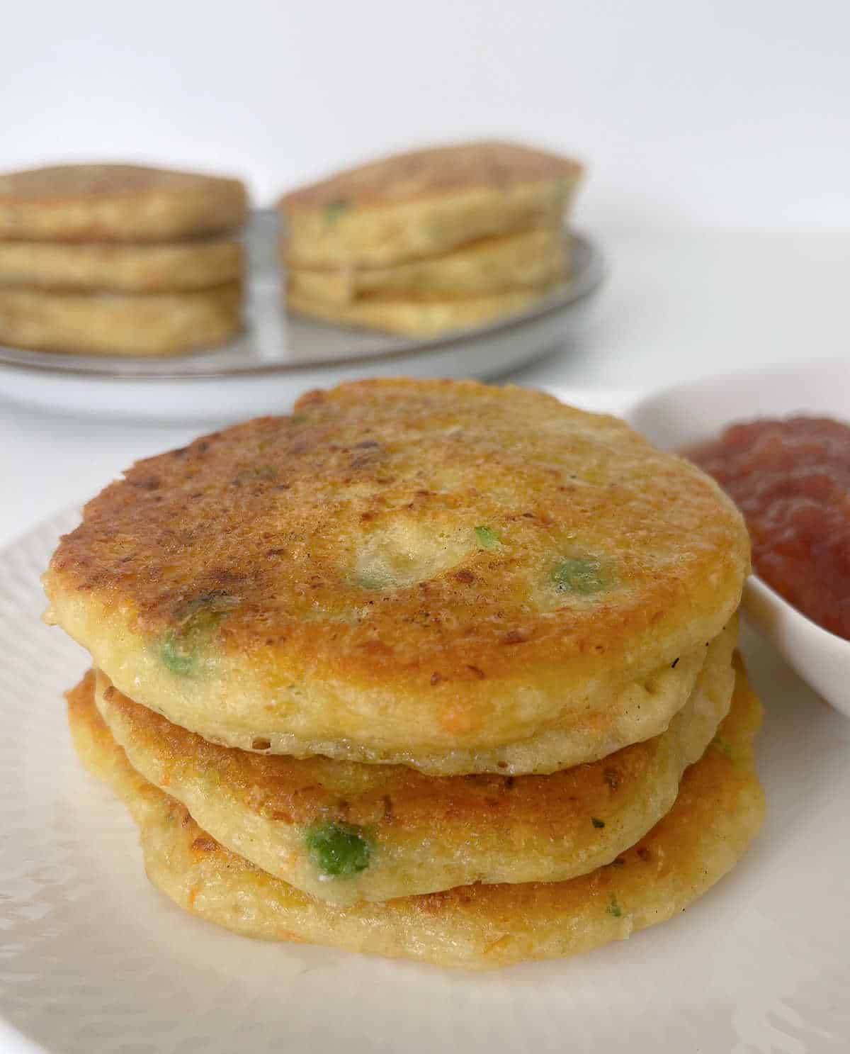 Three vegetable fritters on a white plate. A dish of tomato relish is next to them and a plate with more fritters is in the background.